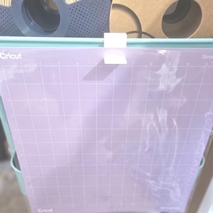 Op de afbeelding: Een lichtpaarse Cricut StrongGrip snijmat met een rasterpatroon, vastgehouden in een turquoise opbergwagen. De mat heeft het Cricut-logo linksonder en de woorden "StrongGrip" rechtsboven.