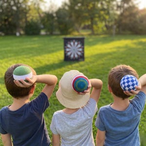 Puede incluir: Tres niños se preparan para lanzar bolas de ganchillo de colores hacia un tablero de dardos en un jardín. Las bolas son verdes, azules y una combinación de rojo, azul y verde. Un niño lleva un sombrero de paja.