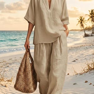 May include: A beige linen outfit, including a V-neck top and wide-leg pants, is worn on a sandy beach. A woven tote bag is carried. The outfit is a neutral color, suitable for warm weather. The background shows the ocean and palm trees.
