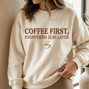 May include: A cream-colored crewneck sweatshirt featuring the brown text COFFEE FIRST, EVERYTHING ELSE LATER printed on the chest, accompanied by a small graphic of a steaming coffee cup with a tiny sparkle icon above it.