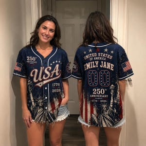 Peut inclure: Un maillot de baseball bleu marine avec des détails blancs, affichant "USA" et "1776-2026". Le devant présente la Statue de la Liberté, et le dos affiche "United States of America" et "Emily Jane" avec le numéro "00".