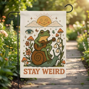 May include: A garden flag with a whimsical design. It features a green frog playing a guitar while riding a snail, with mushrooms, flowers, and a smiling sun. The text "STAY WEIRD" is at the bottom.