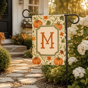 May include: A rectangular garden flag featuring a cream-colored background with a central green-bordered frame containing a large orange letter M. The design includes orange pumpkins and green vines with orange and green autumn leaves throughout.