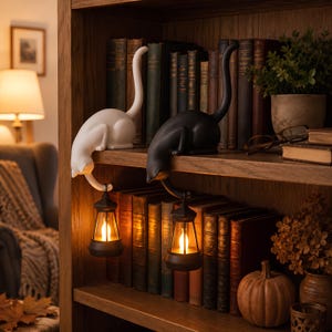 May include: Two cat-shaped ornaments, one white and one black, perched on a wooden shelf. Each cat has a long, curved tail extending downward, holding a small, battery-operated lantern that emits a warm, glowing light. The cats are positioned among various books on the shelf.