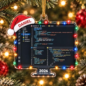 May include: A clear acrylic monitor-shaped Christmas ornament featuring a red Santa hat with the name DANIEL printed on the white trim. The screen displays code, and the base is labeled 2026. Multi-colored LED lights frame the monitor edge.