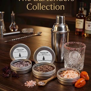 Könnte beinhalten: Die Bartenders Collection. Das Bild zeigt einen Cocktailshaker aus Edelstahl, ein Kristallglas mit gezuckertem Rand und Eis sowie kleine Dosen mit aromatisierten Salzen. Die Salze umfassen Hibiskus- und Espresso-Sorten. Ein Holzlöffel und Paprika sind ebenfalls vorhanden.