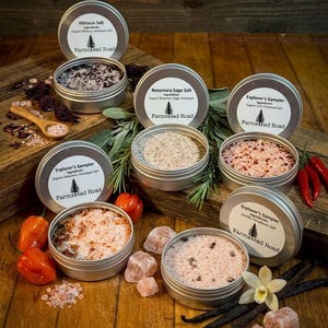 Könnte beinhalten: Eine Auswahl kleiner, runder Metalldosen, gefüllt mit verschiedenen aromatisierten Salzen. Jede Dose hat ein Etikett mit dem Namen des Salzes und der Marke "Farmslead Road". Zu den Zutaten gehören Hibiskus, Rosmarinsalbei, Habanero, Vanille und Himalaya-Salz. Rote Paprika und Vanilleschoten sind ebenfalls zu sehen.