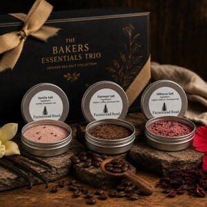 May include: A gift set featuring three tins of infused sea salt: Vanilla, Espresso, and Hibiscus. The tins are displayed with coffee beans, a wooden spoon, and a decorative box with a bow. The text on the box reads "The Bakers Essentials Trio".