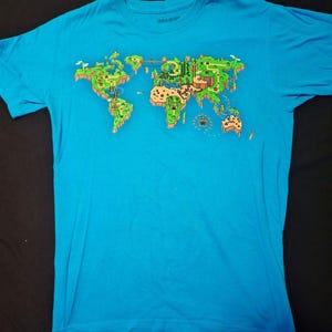 May include: A turquoise t-shirt featuring a colorful world map design in a video game style. The map includes green landmasses, brown terrain, and various game elements. The shirt is made of a soft material.