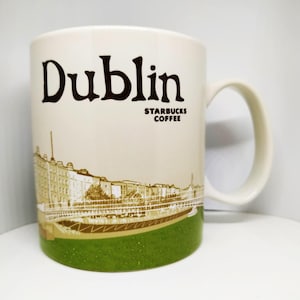 May include: Cream-colored Starbucks mug featuring the city name "Dublin" in dark brown lettering. The mug has a scenic illustration of Dublin's architecture and a green textured base. The Starbucks Coffee logo is also visible.