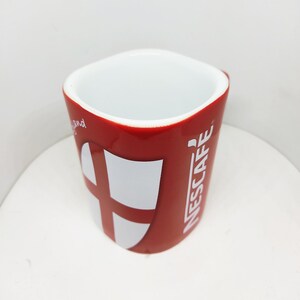 May include: A red ceramic mug with a white interior featuring the Nescafé logo in white vertical text and a white shield graphic containing a red cross. The mug has a rounded square shape and is viewed from a high angle against a plain background.