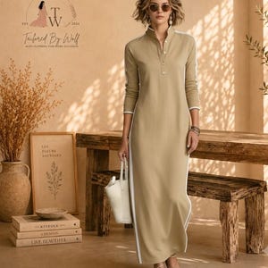 May include: A long-sleeved, ankle-length beige polo dress featuring a button-up placket, a stand-up collar, and white vertical stripes running down the sides of the sleeves and the length of the dress, paired with a white handbag and sandals.