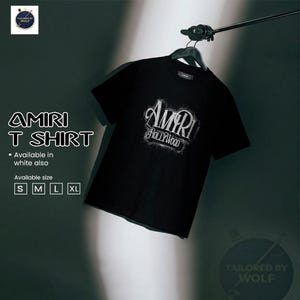 Könnte beinhalten: Schwarzes T-Shirt mit dem Text "AMIRI HOLLYWOOD" in weißer, stilisierter Schrift. Das Shirt hängt an einem schwarzen Kleiderbügel. Der Text "AMIRI T SHIRT" wird links mit Größenoptionen angezeigt.