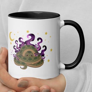 May include: A white ceramic mug with a black rim and handle featuring a graphic illustration of a coiled green snake surrounded by purple swirling smoke, a yellow crescent moon, and small yellow stars against a plain white background.