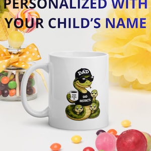 Peut inclure: Mug en céramique blanche avec un serpent de dessin animé portant un chapeau "DAD" et des lunettes de soleil, tenant une tasse de café. Le mug porte le texte "DAD INSTINCTS" et "DAD FUEL". Le mug est personnalisé avec le texte "PERSONNALISÉ AVEC LE NOM DE VOTRE ENFANT".