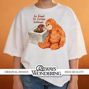 Op de afbeelding: Wit T-shirt met een aquarel illustratie van twee orang-oetans. Eén zit buiten een betonnen doos en kijkt naar een kleinere orang-oetan binnenin. De tekst "be kind, be brave, believe" staat boven de illustratie. Het shirt heeft ook de tekst "Always Wondering".
