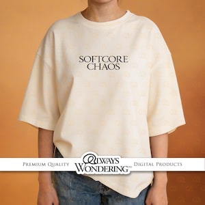 Pode incluir: Camiseta creme com a frase "SOFTCORE CHAOS" em preto no centro. A camiseta de manga curta tem um corte folgado. Uma faixa branca na parte inferior mostra "ALWAYS WONDERING" e "DIGITAL PRODUCTS".