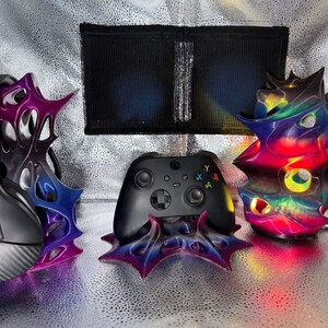 May include: A black gaming controller and black headphones resting on three 3D-printed stands with a gradient colour scheme of purple, blue, and pink. The stands feature an organic, web-like structure with pointed protrusions and smooth curves.