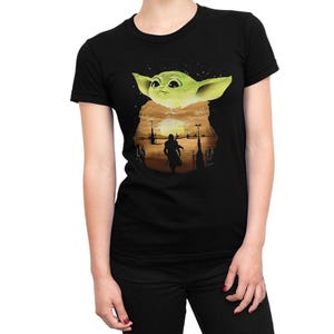 Pu&ograve; includere: T-shirt nero con grafica di Baby Yoda verde sopra una scena desertica con una silhouette. Il design include un tramonto e degli edifici. La maglietta ha girocollo e maniche corte.