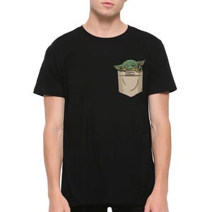 May include: Black t-shirt featuring a pocket with a graphic of Grogu, also known as Baby Yoda, peeking out. The pocket is tan, and Grogu is green with large eyes. The t-shirt is a short-sleeved crew neck.