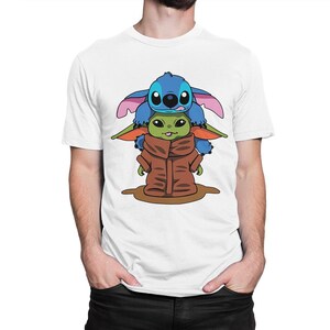 May include: White t-shirt with a cartoon graphic of Stitch from Lilo & Stitch and Grogu (Baby Yoda) from The Mandalorian. Stitch is blue with large ears, and Grogu is green and wearing a brown cloak. The characters are stacked on top of each other.