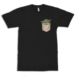 May include: Black t-shirt featuring a pocket with a green cartoon character peeking out. The character has large eyes and pointed ears. The pocket is tan and has a small rectangular detail.