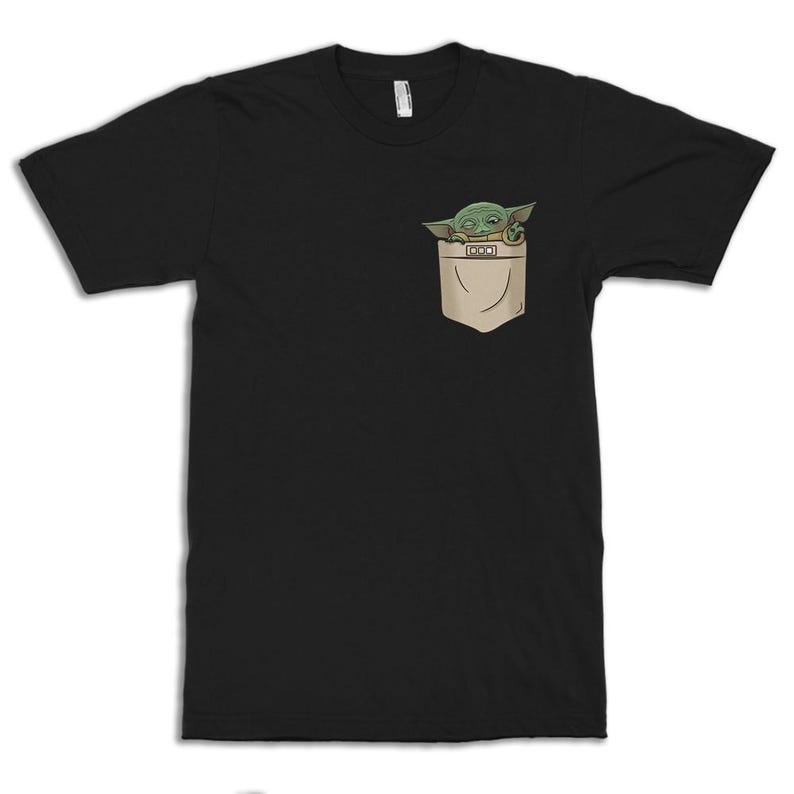 May include: Black t-shirt featuring a pocket with a green cartoon character peeking out. The character has large eyes and pointed ears. The pocket is tan and has a small rectangular detail.