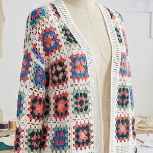 Può includere: Un cardigan all'uncinetto bianco con davanti aperto e un colorato motivo a piastrelle della nonna. I quadrati presentano sfumature di rosso, blu e verde. Il cardigan è esposto su un manichino, mostrando il suo design aperto e l'intricato lavoro a maglia.