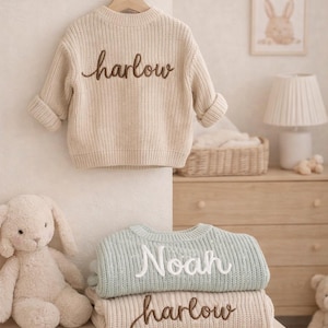 May include: A stack of children's sweaters in cream and light blue, with embroidered names. The top sweater is cream with "harlow" in brown script. The next sweater is light blue with "Noah" in white script, and the bottom sweater is cream with "harlow" in brown script.