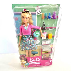Peut inclure: Une poupée Barbie aux cheveux blonds portant un haut rose à manches longues, une jupe à motifs floraux et des lunettes noires. L'emballage contient un globe, un microscope, un ordinateur portable, des éprouvettes et des livres. Texte Barbie You Can Be Anything.