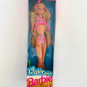 Op de afbeelding: Een vintage Jazzie Glitter Beach Barbie pop in de originele verpakking. De pop heeft lang blond haar en draagt een roze en bloemenprint bikini. De doos is roze en blauw met de tekst "Glitter Beach Barbie" op de voorkant.