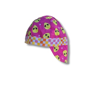 May include: A pink welding cap with a repeating pattern of yellow alien faces and small white stars. The cap features a checkered band in orange, white, and yellow. The cap is designed to protect the head and neck.