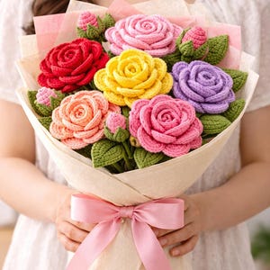 Puede incluir: Un ramo de rosas de ganchillo en tonos rojo, amarillo, morado, rosa y melocotón, acentuado con hojas y capullos de ganchillo verdes. Las flores están envueltas en papel beige claro y atadas con un lazo de cinta de satén rosa claro.