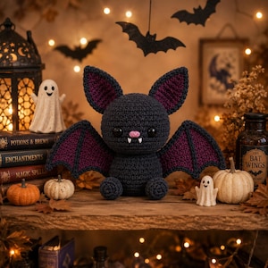Può includere: Un peluche a forma di pipistrello lavorato all'uncinetto in grigio scuro con grandi orecchie e ali interne viola, occhi a bottone neri, un piccolo naso rosa e due zanne bianche. Il pipistrello è centrato e ha un corpo rotondo e testurizzato.