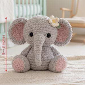 May include: A handmade gray crochet elephant, approximately 8 inches tall, with pink inner ears and foot pads. It features black safety eyes and a small flower on its head. The elephant is sitting on a cream-colored rug.