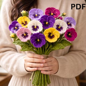 以下が含まれることがあります： 紫、ピンク、黄色、白など、様々な色の手編みのパンジーのブーケ。花は緑の葉と茎でまとめられ、ひもで結ばれています。画像には「MOTHERS」と「DAY GIFT」という文字が含まれています。