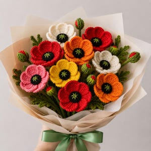 May include: A bouquet of crocheted flowers featuring red, orange, yellow, pink, and white poppy-like blossoms with black centres and green stems. The arrangement includes several small green buds and textured leaves, wrapped in light beige paper and tied with a green ribbon.