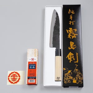 May include: A Japanese kitchen knife with a dark, hammered steel blade and a light wood handle, presented in a black box with gold Japanese text. Beside it is a rectangular sharpening stone with a label and a small circular red and white sticker.
