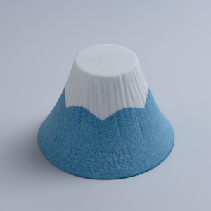Op de afbeelding: Een keramische sculptuur van de berg Fuji. De berg heeft een blauwe basis en een witte, met sneeuw bedekte top. De sculptuur is kegelvormig met een textuur, tegen een effen achtergrond.