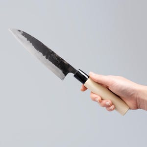 May include: A Japanese-style kitchen knife with a dark, textured forged steel blade featuring visible kanji characters and a sharp, pointed tip. The handle is made of light-colored, smooth wood with a black ferrule connecting it to the blade.