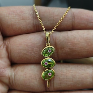 Könnte beinhalten: Eine Goldkette mit einem vertikalen Anhänger, der aus drei ovalen, grünen Peridot-Steinen besteht, in deren Mitte jeweils ein kleiner, runder, rosafarbener Edelstein eingefasst ist. Der Anhänger hängt an einer feinen Goldkette in linearer Form.