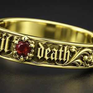 May include: A gold ring featuring an intricate scrollwork pattern and the engraved text 'til death' in a gothic font. A single round, deep red gemstone is set within a decorative metal frame between the words on the band's outer surface.