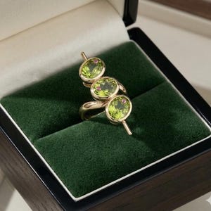 Puede incluir: Un anillo de oro con tres piedras preciosas de peridoto de color verde claro y corte ovalado dispuestas en fila vertical. Cada peridoto está acentuado con una pequeña piedra preciosa redonda de color rosa intenso engastada en la piedra.