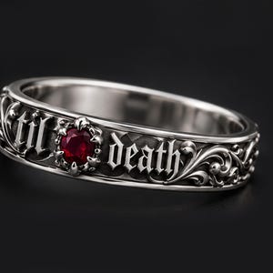 Könnte beinhalten: Ein silberfarbener Metallring mit einem komplizierten Schnörkelmuster und dem eingravierten Text 'til death' in einer gotischen Schriftart. Ein kleiner, runder, tiefroter Edelstein ist in die Mitte des Bandes eingefasst und von verzierten Metalldetails umgeben.