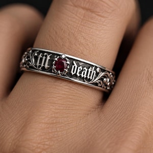 Peut inclure: Une bague en argent ornée d'un motif floral complexe et d'une pierre précieuse rouge foncé en son centre. La surface métallique est gravée du texte 'til death' dans une police gothique, avec des lettres mises en valeur par une finition oxydée.