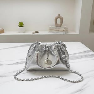 Puede incluir: Un bolso plateado con la parte superior fruncida y una correa de cadena. El bolso tiene un pequeño dije redondo con una perla. El bolso está hecho de un material brillante y metálico.