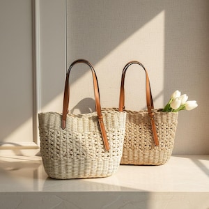 Könnte beinhalten: Zwei gewebte Tragetaschen mit braunen Ledergriffen. Eine Tasche enthält weiße Tulpen. Die Taschen bestehen aus einem hellbeigen Material mit einem gewebten Muster. Die Griffe sind mit braunen Lederriemen befestigt.