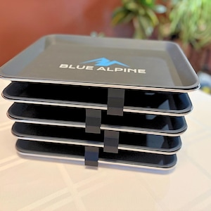 May include: A stack of four black trays with silver trim, held together by black clips. The top tray has the words "BLUE ALPINE" in white with a blue mountain logo.