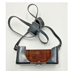 May include: A vintage black leather camera case with a matching strap. The case has a rectangular shape with a brown interior and a silver-colored clasp. A small, round, zippered case is attached to the strap.