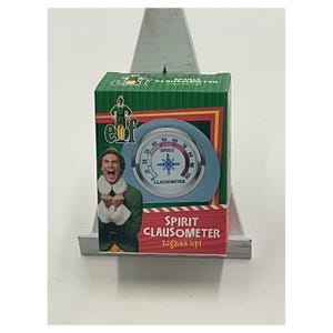 May include: A novelty "Spirit Clausometer" from the movie Elf. The box is green with a picture of Buddy the Elf. The Clausometer has a blue face with a needle and a snowflake design. The text "Spirit Clausometer Lights up!" is displayed on the box.